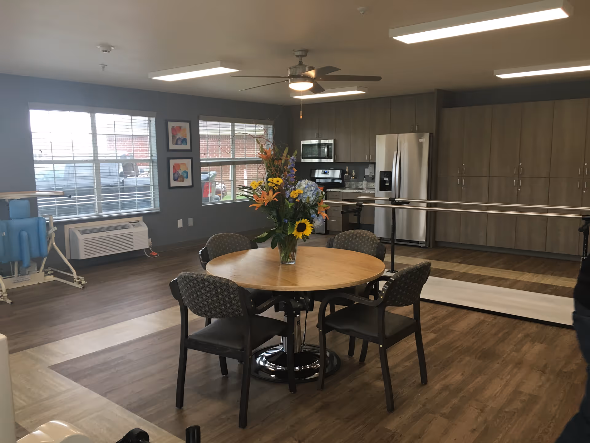Round wooden dining table with chairs, floral centerpiece, and kitchenette in a communal lounge.