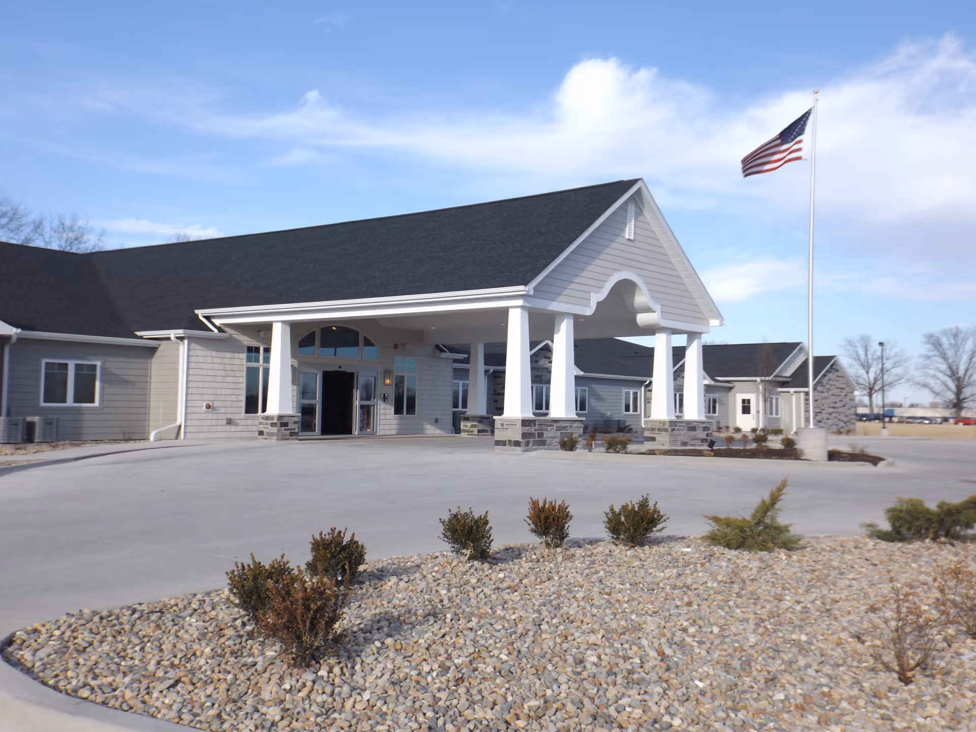 Front exterior of a single-story memory care facility with a covered entrance/porte-cochère, American flag on a pole, and gravel landscaping.