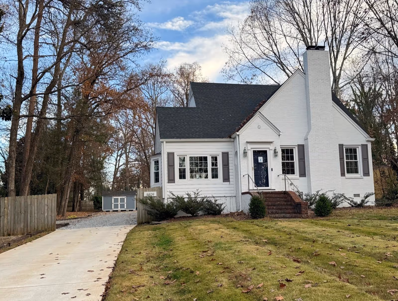 White suburban house with blue front door, gray shutters, brick steps, driveway, and surrounding trees.