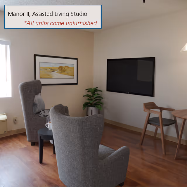 A small assisted living studio room with two gray upholstered armchairs facing a wall-mounted flat screen TV. There is a small round black table between the chairs with a white cup on it. A framed landscape painting hangs on the wall near a window letting in natural light. A potted plant is placed in the corner, and a wooden chair and round table are visible on the right side. The floor is wooden, and the walls are painted light beige. A sign on the wall reads 'Manor II, Assisted Living Studio *All units come unfurnished.'