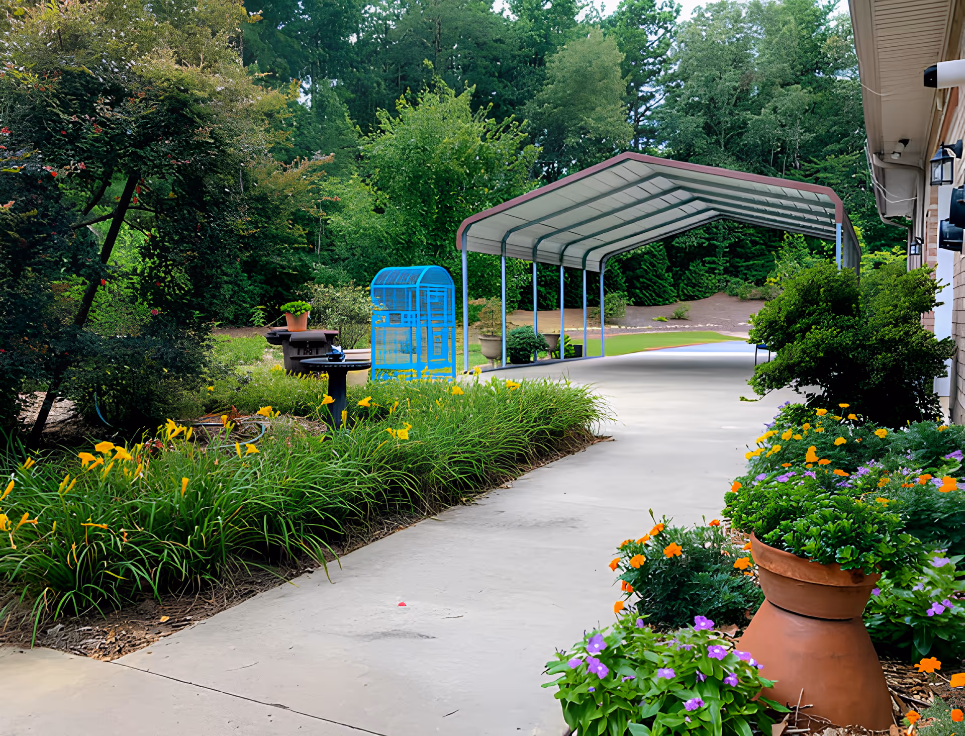 A paved outdoor walkway lined with colorful flowers and green plants on both sides. There is a blue birdcage, a birdbath, and a covered metal carport structure in the background. Trees and bushes surround the area, creating a lush garden setting.