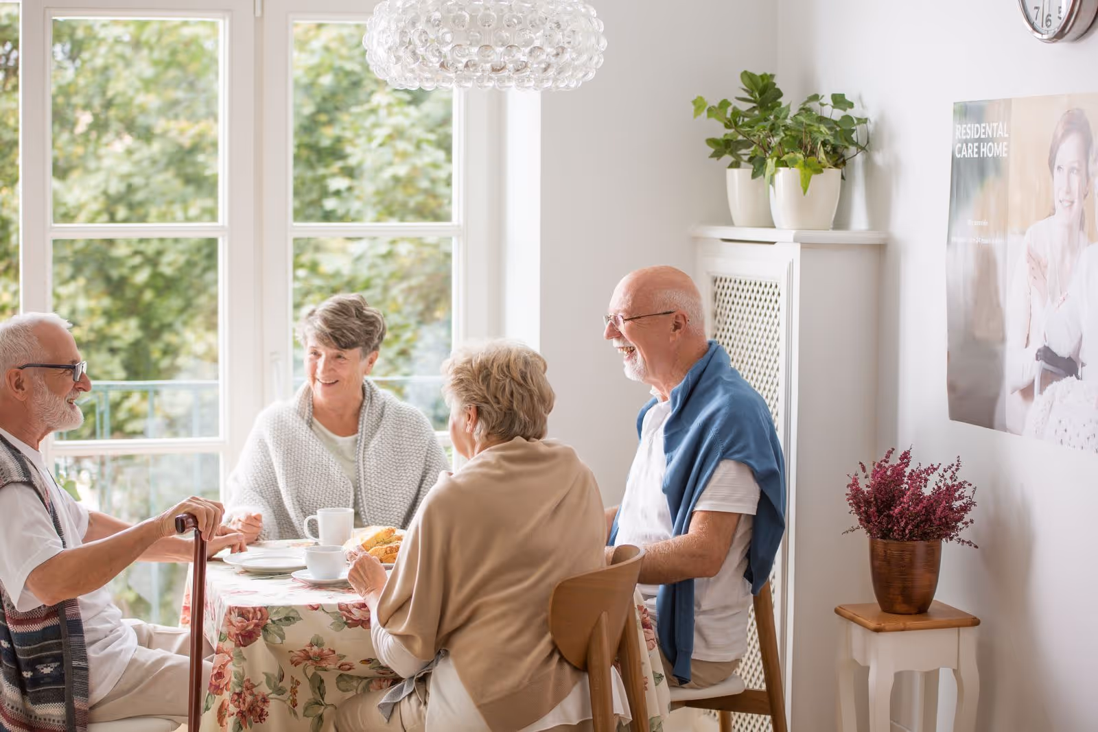 Four older adults seated around a floral-covered dining table in a bright room with large windows, enjoying tea and conversation.