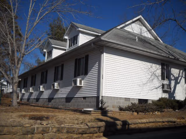 White, multi-story building with pitched roof and window air conditioning units along a stone foundation.
