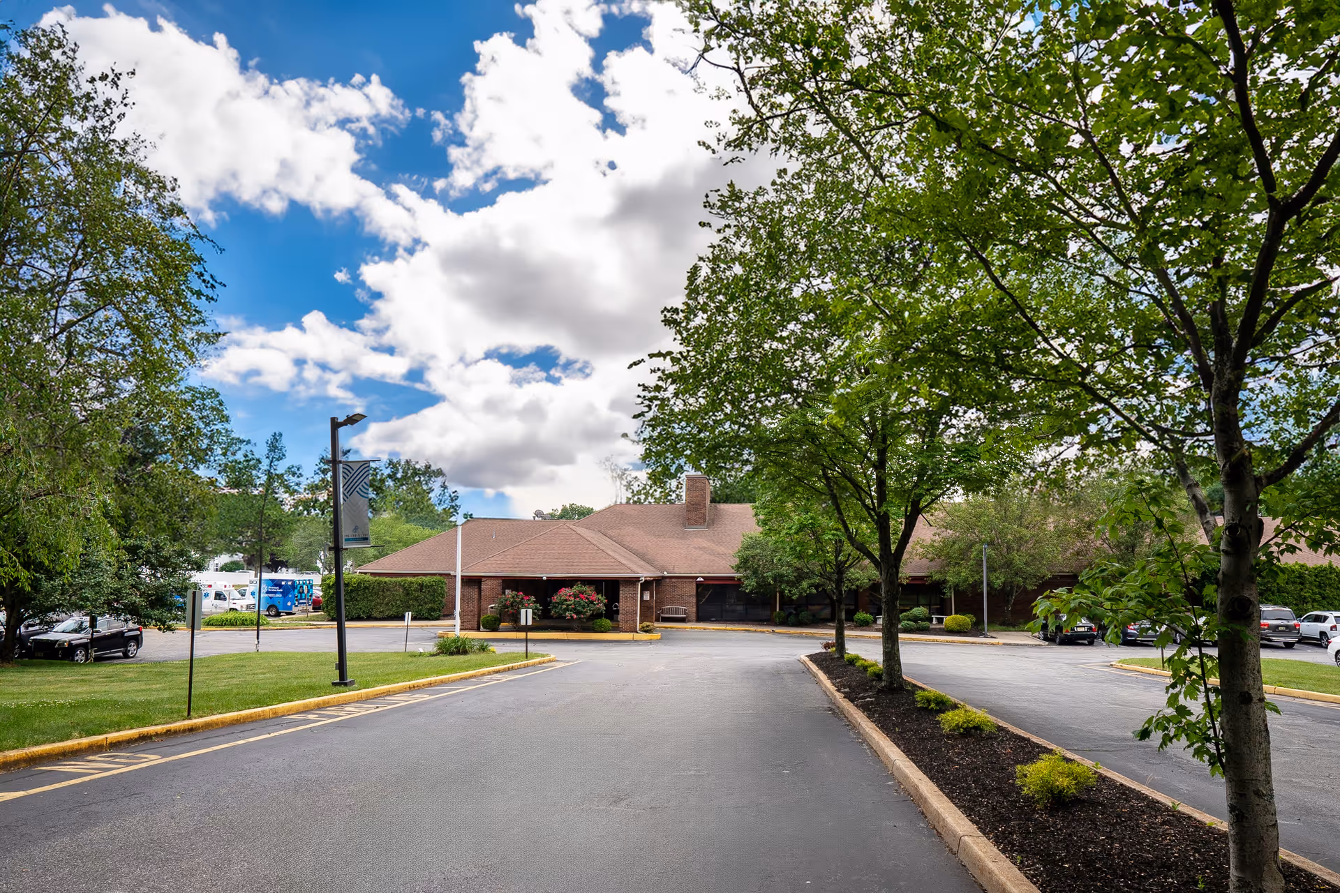 View of the front exterior of a single-story brick building with a brown roof, surrounded by trees and a parking lot under a partly cloudy blue sky.