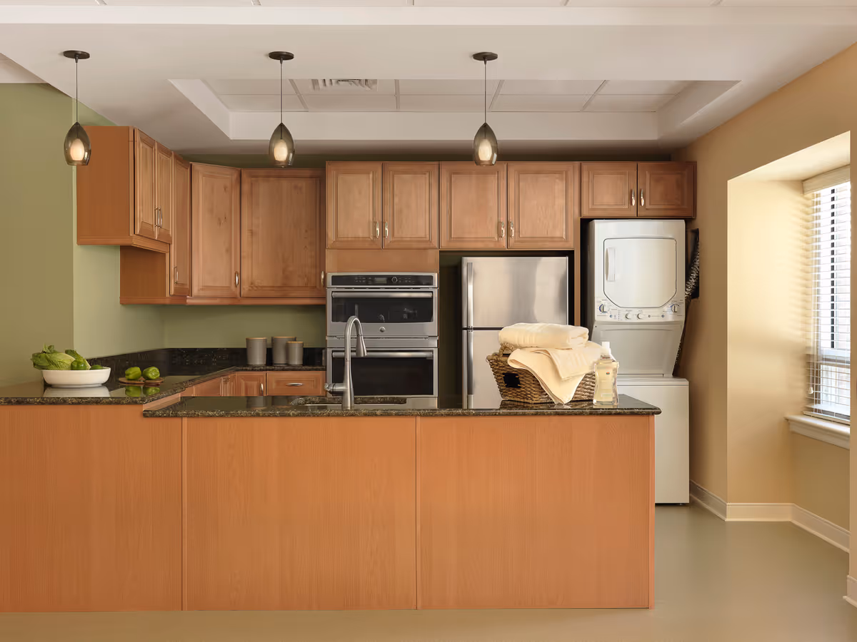 A modern kitchen with wooden cabinets, a stainless steel refrigerator, built-in oven, and a stacked washer and dryer unit. The kitchen features a granite countertop island with a sink and a basket containing folded towels and a bottle of soap. There are three pendant lights hanging from the ceiling and a window with blinds on the right side.