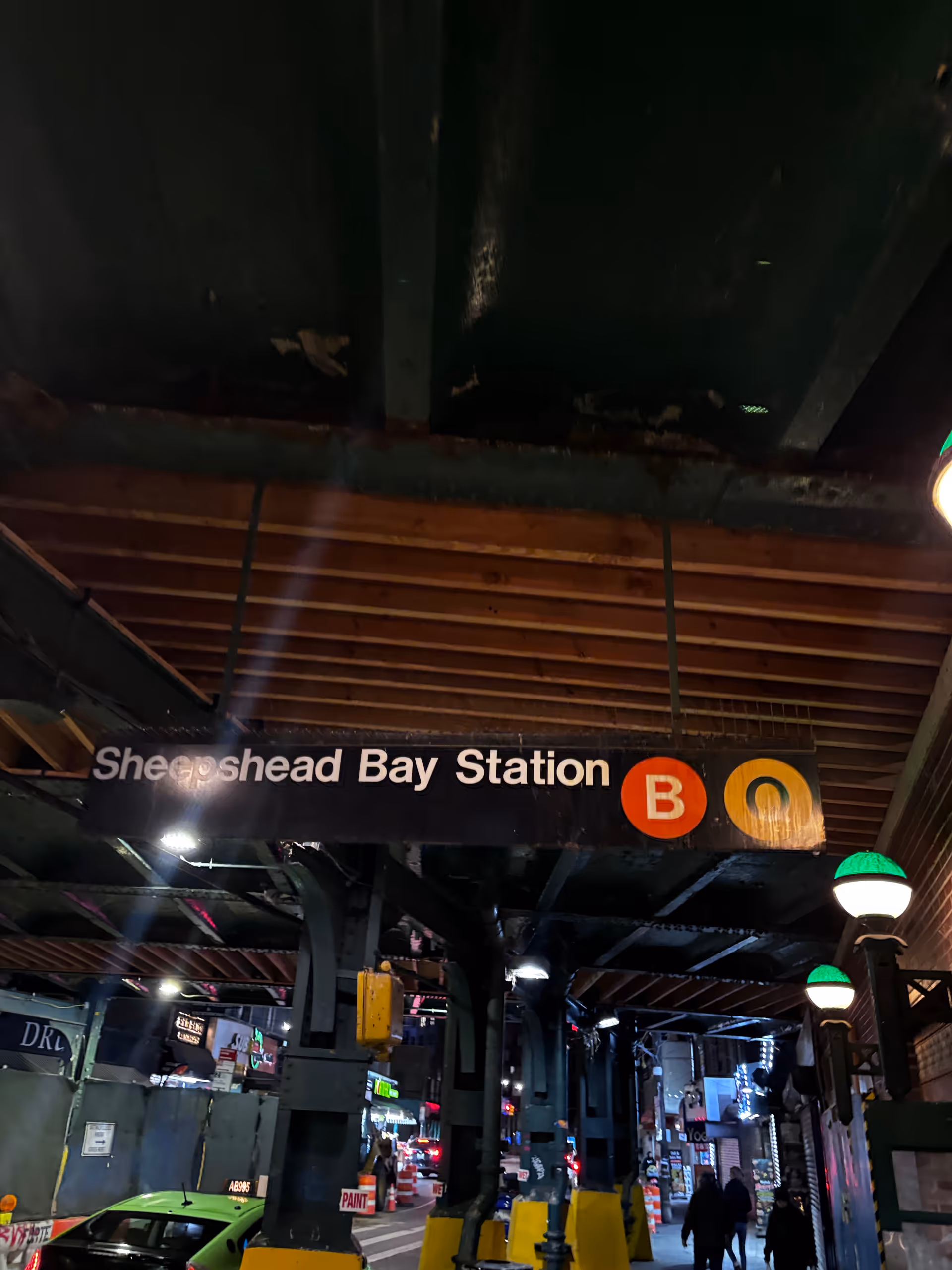 Dark subway station interior with Sheepshead Bay Station sign, B and Q logos, and pedestrians.
