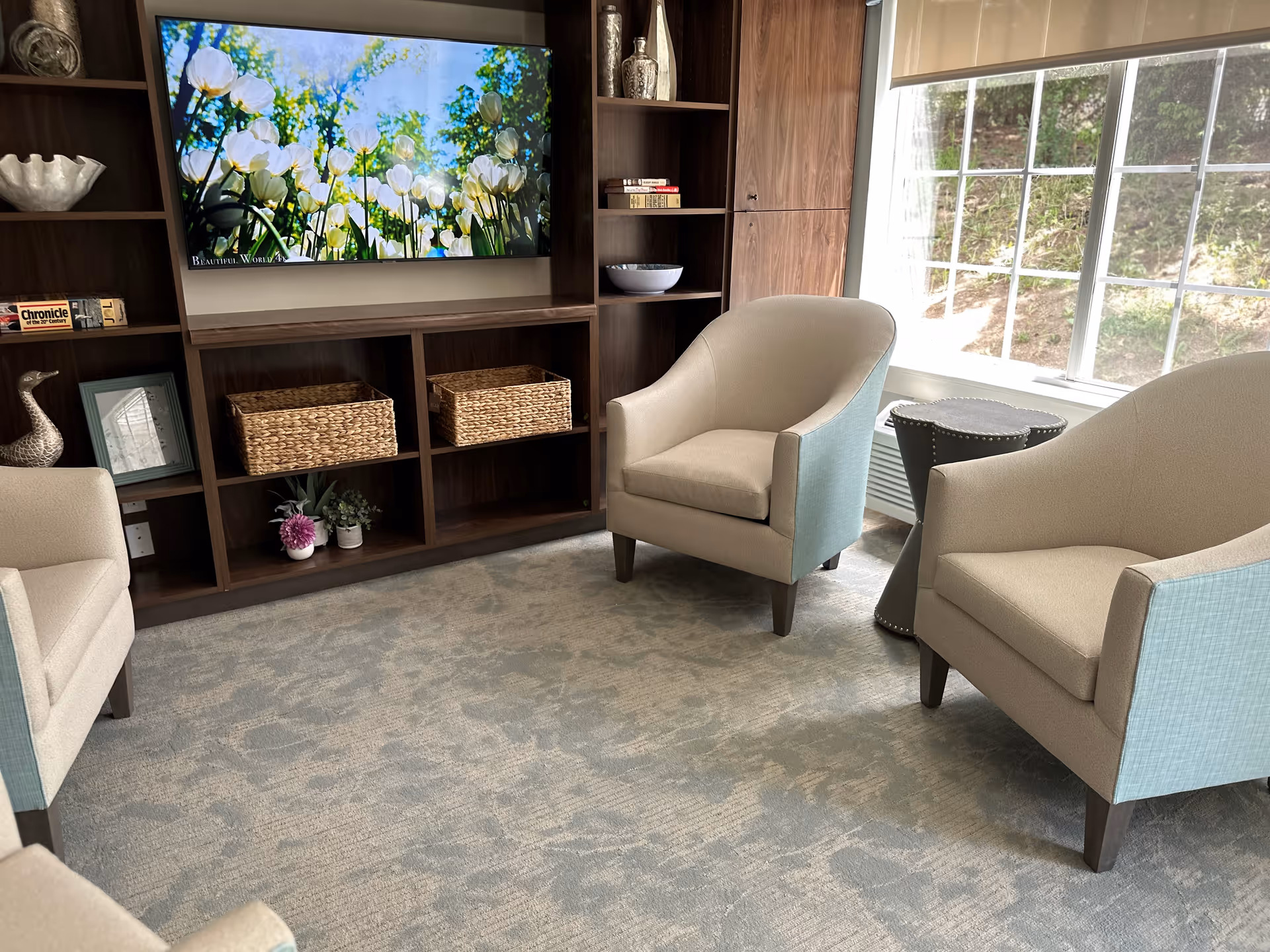 A cozy living room area with three beige armchairs arranged around a small round side table. Behind the chairs is a large wooden shelving unit with decorative items, baskets, and a flat-screen TV displaying an image of white tulips and greenery. A large window lets in natural light, illuminating the room.