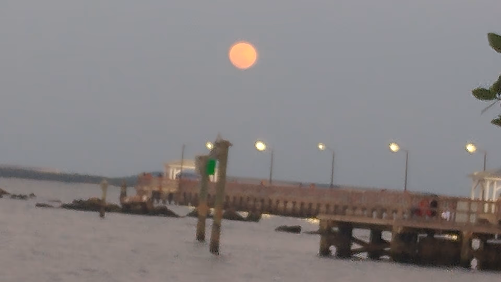 Orange full moon above a wooden pier extending over calm water at dusk with lamps.