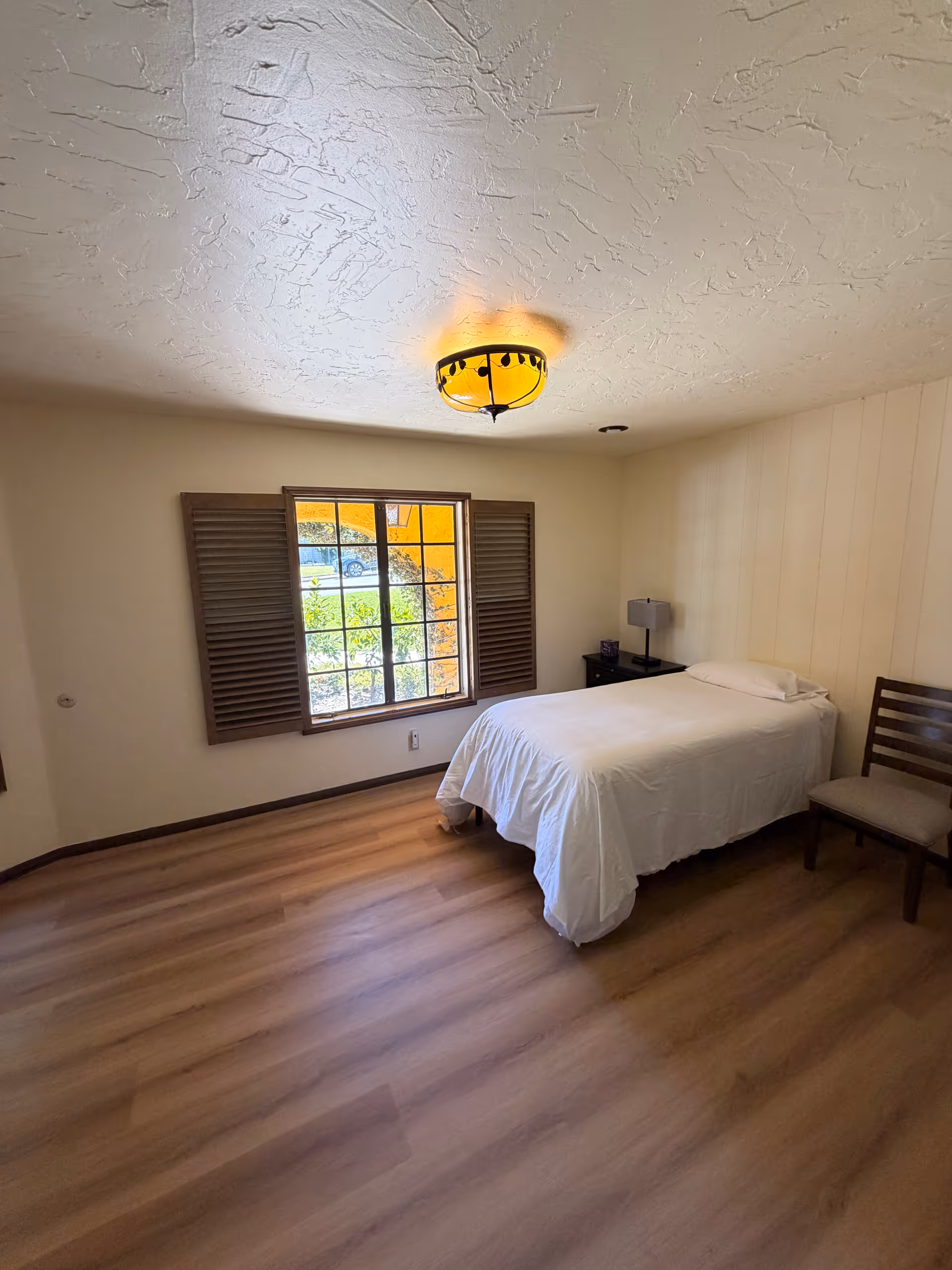 Simple bedroom with bed, window, nightstand, lamp, chair, and wood flooring.