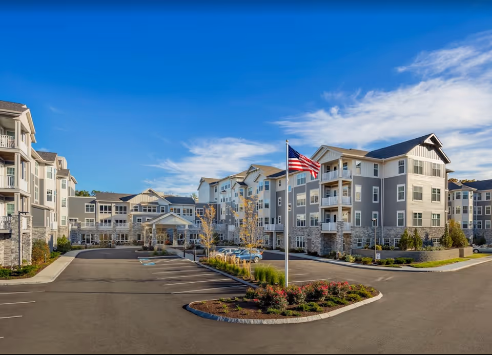 Front exterior of a multi-story senior living building with balconies, an American flag, and a parking area under a blue sky.