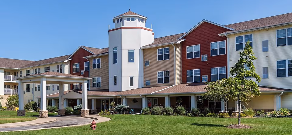 Front exterior of a three-story senior living building with a covered entrance, tower feature, and green lawn under a clear blue sky.