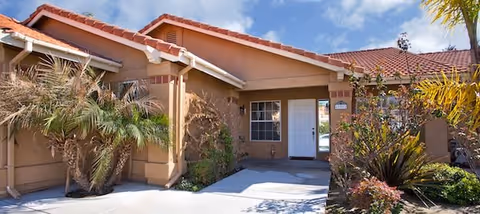 Front entrance of a single-story tan stucco building with a red tile roof, white door, and landscaped plants.