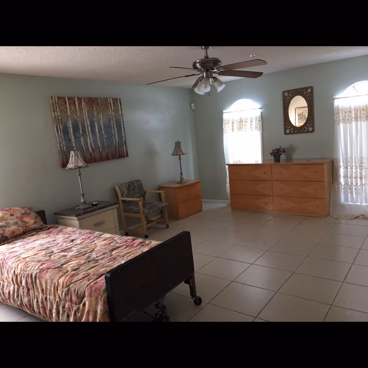 A sparsely furnished bedroom with a single bed, dresser, chair, bedside tables and lamps, ceiling fan, and tiled floor.