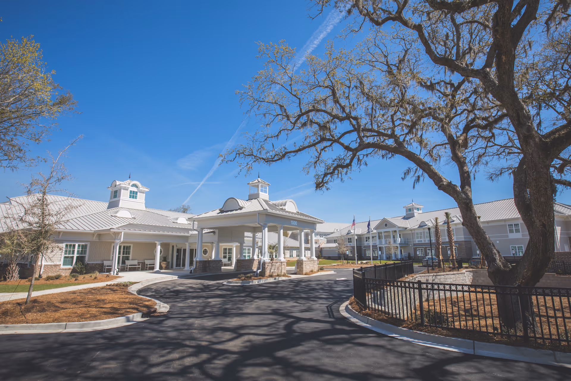 Exterior view of Wellmore of Daniel Island senior living facility on a clear sunny day, showing a large driveway, entrance canopy, multiple buildings with white roofs, and a large tree with bare branches in the foreground.