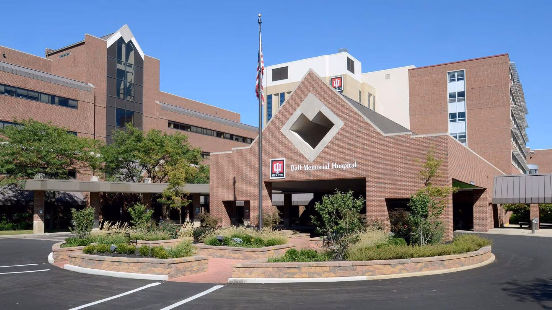 Brick hospital building with circular driveway, landscaped grounds, flag, and IU Health Ball Memorial Hospital