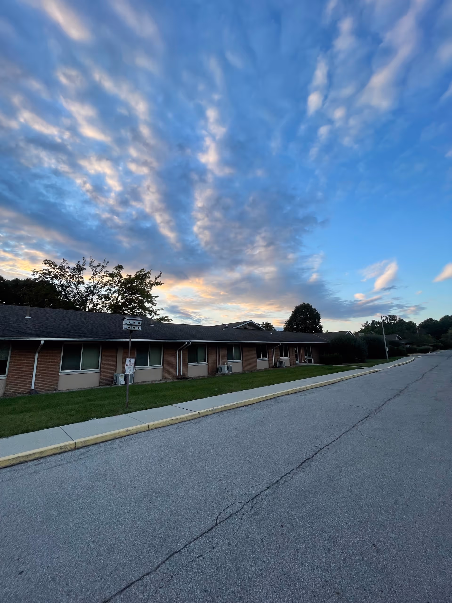 Sunset sky above a single-story brick senior living building along a quiet street.