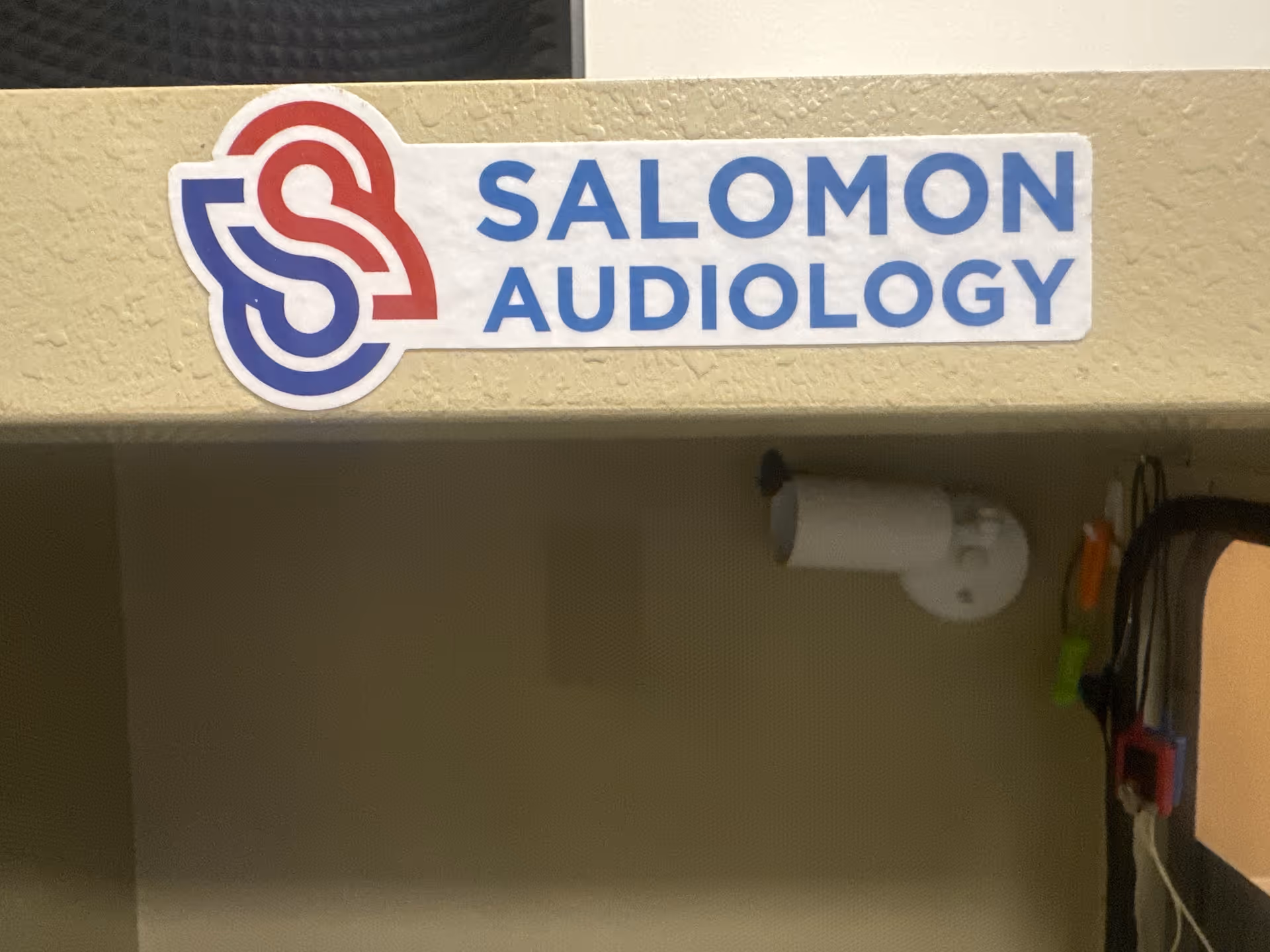 Sign reading Salomon Audiology mounted on a beige wall, with a small camera nearby.