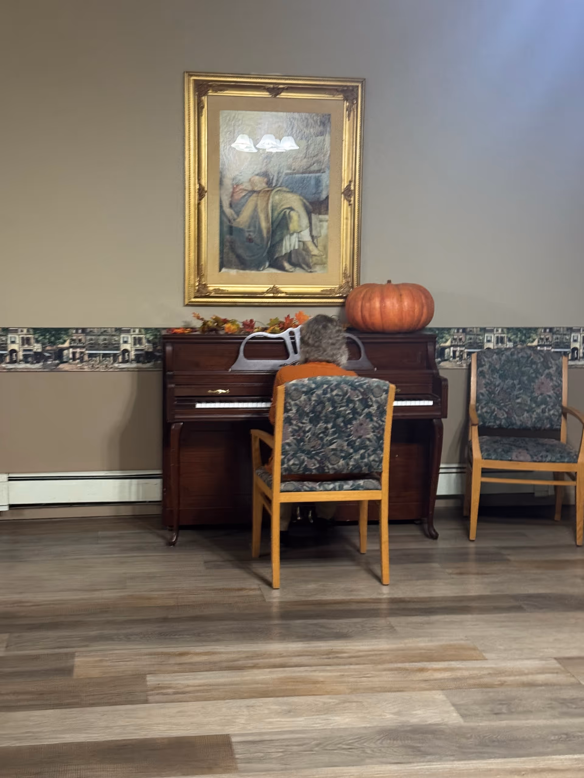 An elderly resident sits at a wooden piano in a patterned lounge.