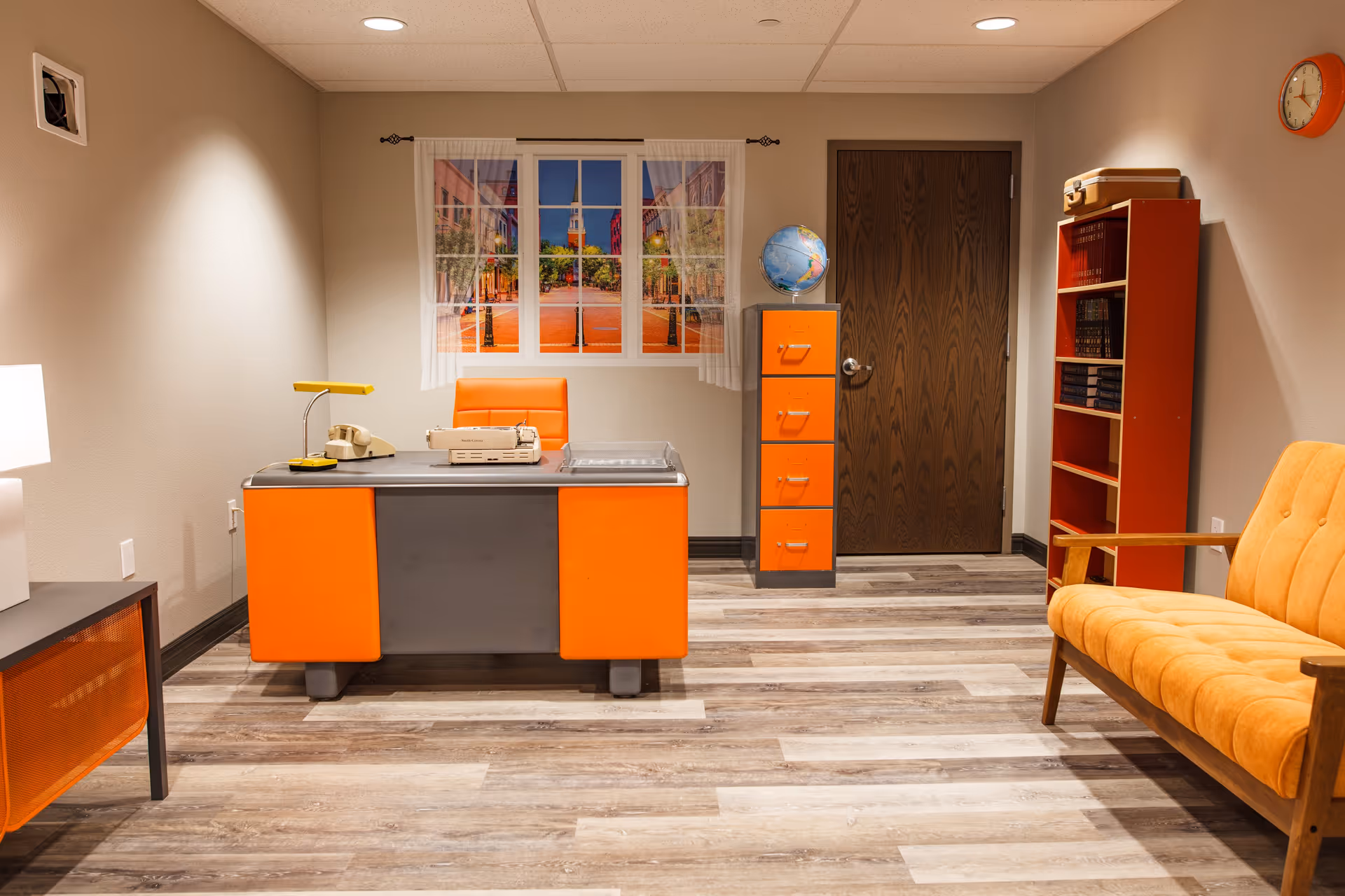 An office room with a retro style featuring an orange and gray desk with a vintage telephone and typewriter on top. Behind the desk is an orange chair, a window with a scenic city street view curtain, and a globe on an orange filing cabinet. To the right, there is a wooden bookshelf with books and a small suitcase on top, and an orange cushioned armchair. The room has wood-patterned flooring and beige walls with a clock on the upper right wall.