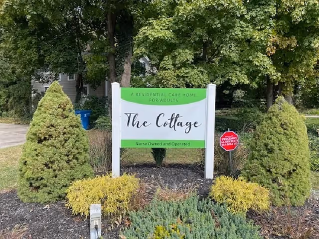 Front yard sign for The Cottage residential care home with trimmed evergreens