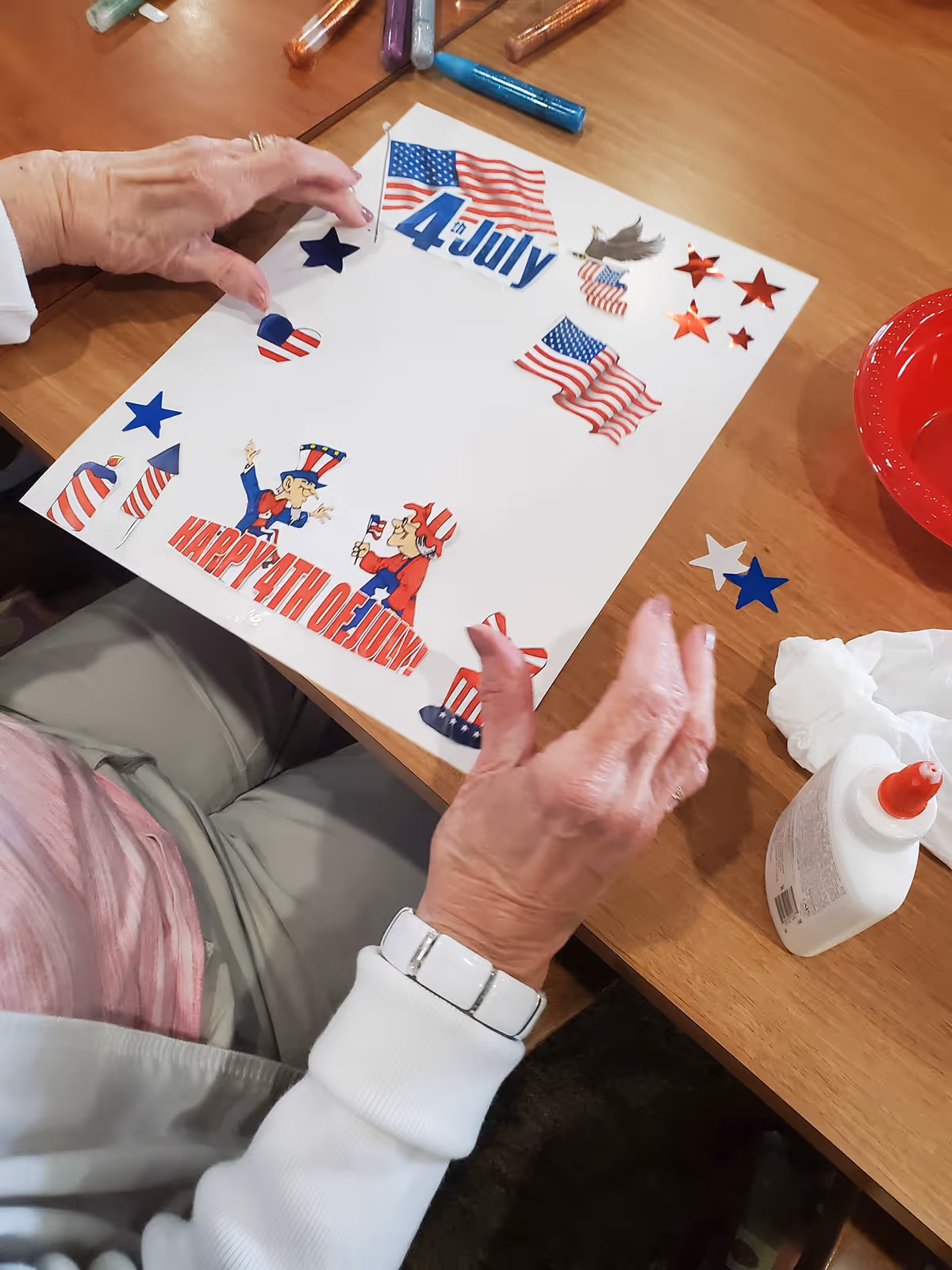 An elderly person sitting at a wooden table is creating a 4th of July themed craft. The craft features patriotic decorations including American flags, stars, fireworks, and cartoon characters dressed in red, white, and blue. Various craft supplies like glue and glitter tubes are visible on the table.