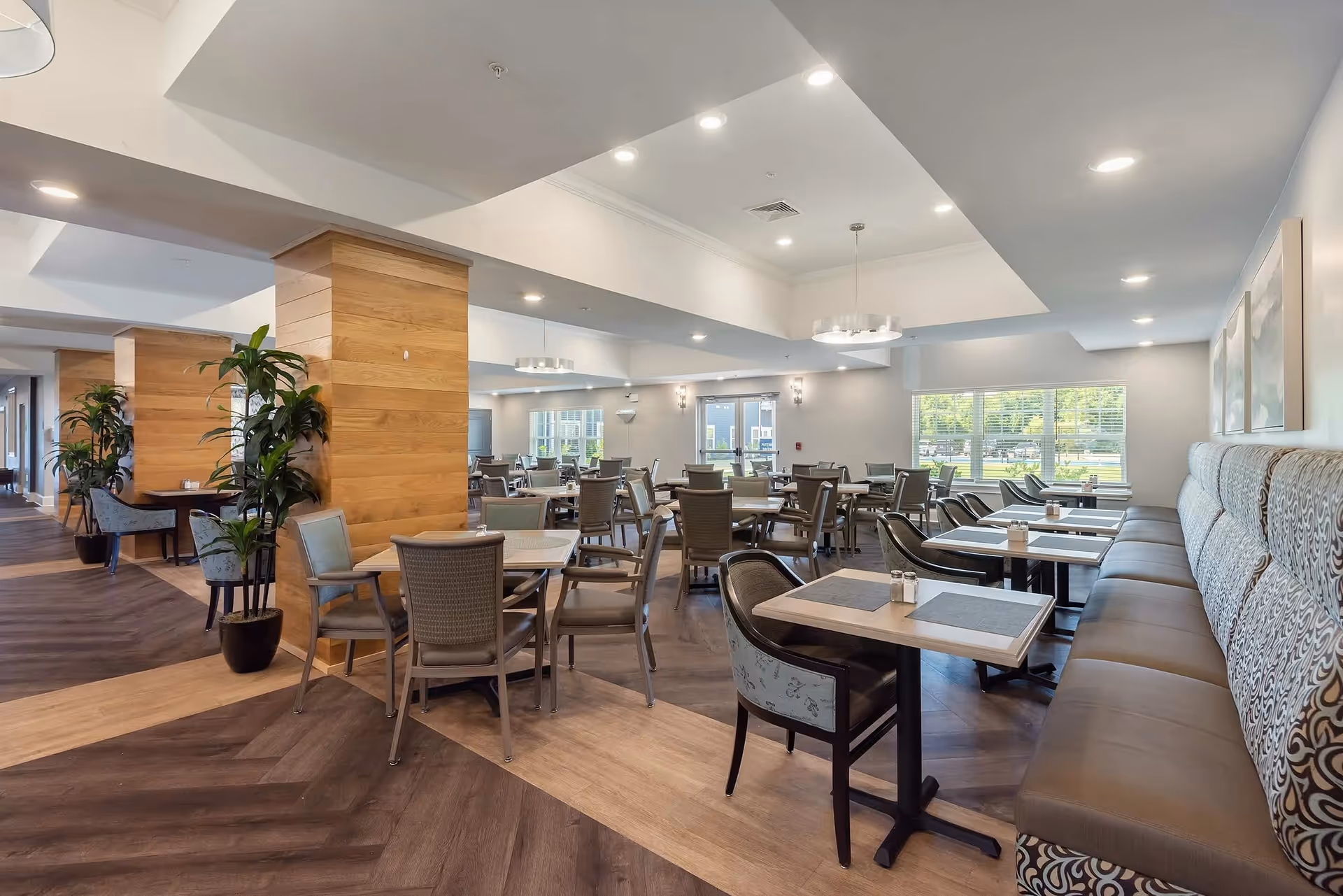 A spacious and well-lit dining area with multiple tables and chairs arranged neatly. The room features a combination of wooden and upholstered seating, with large windows allowing natural light to fill the space. The floor has a patterned wood design, and there are several plants and modern light fixtures enhancing the ambiance.