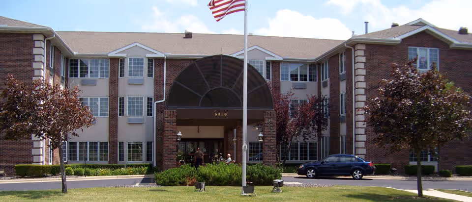Front exterior view of a three-story brick building with multiple windows, a covered entrance with an arched roof, an American flag on a flagpole in front, and a parked car to the right. There are small trees and shrubs in the landscaped area around the entrance.