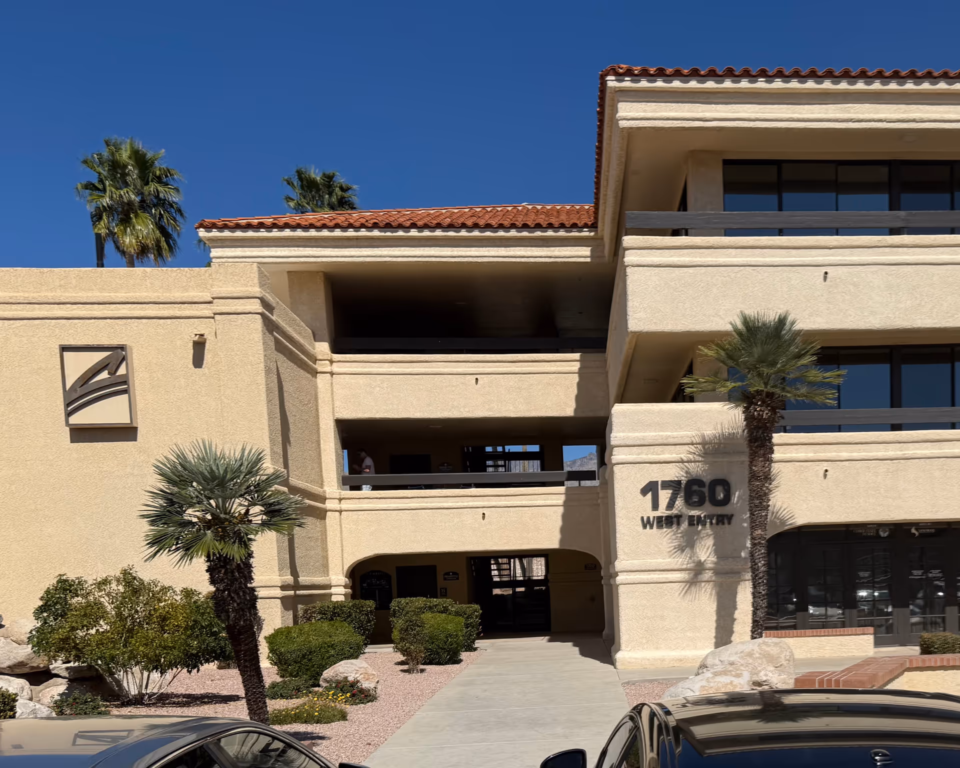 Exterior view of a beige multi-level senior facility with palm trees and parked cars.