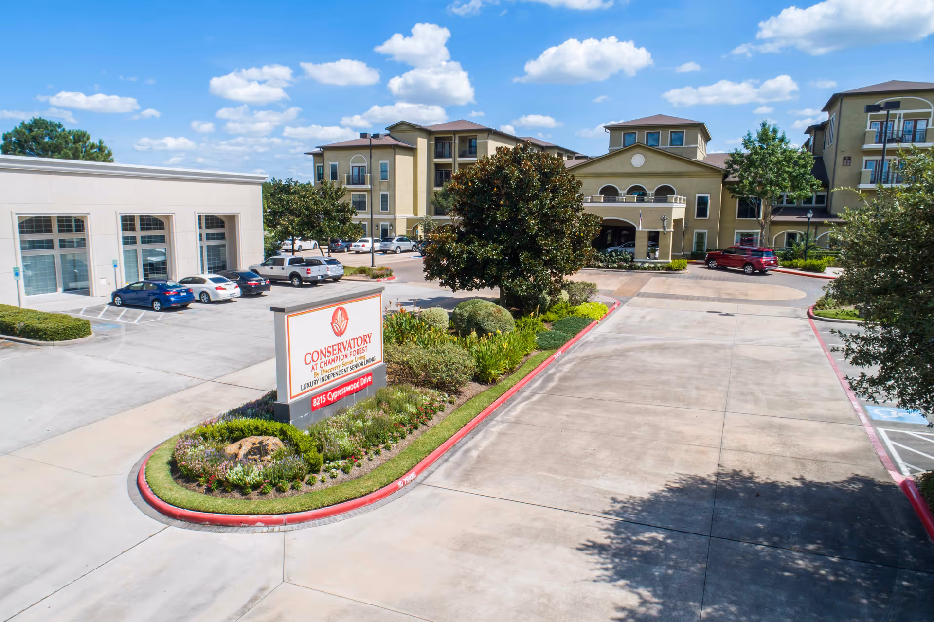 Exterior view of Conservatory At Champion Forest senior living facility showing the main entrance, parking area with several cars, landscaped greenery, and a clear blue sky with clouds.