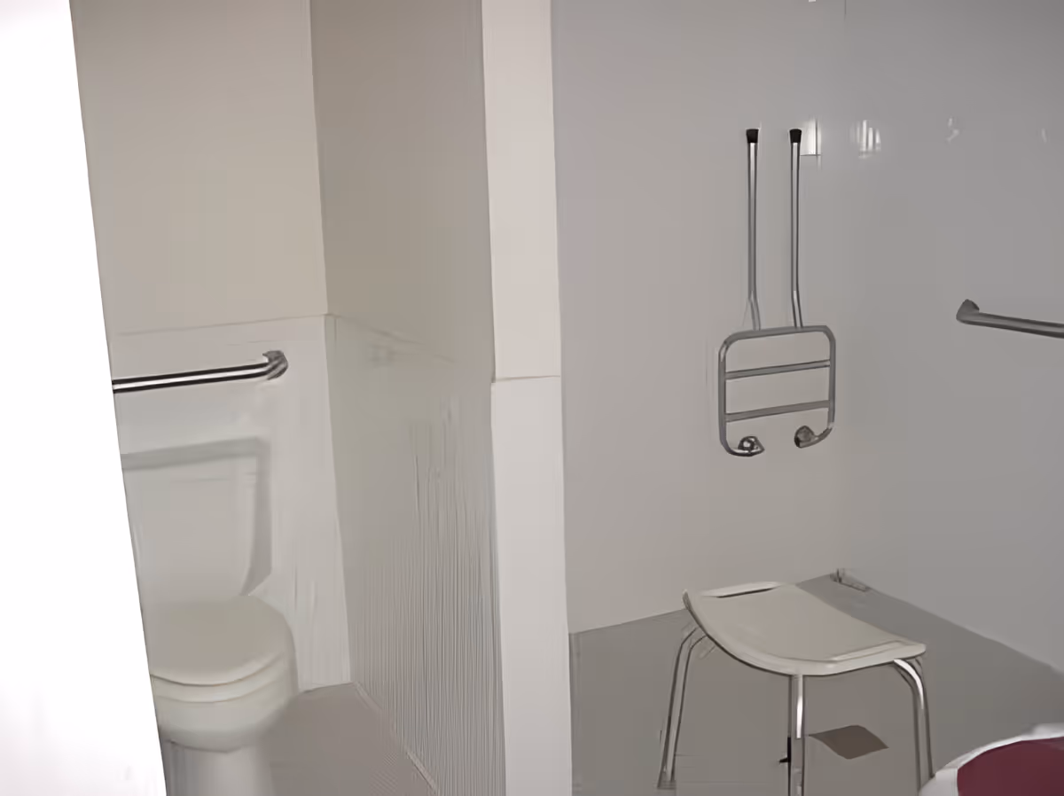 A bathroom with a white toilet, grab bars on the walls, a white shower chair, and a foldable shower seat mounted on the wall, designed for accessibility.