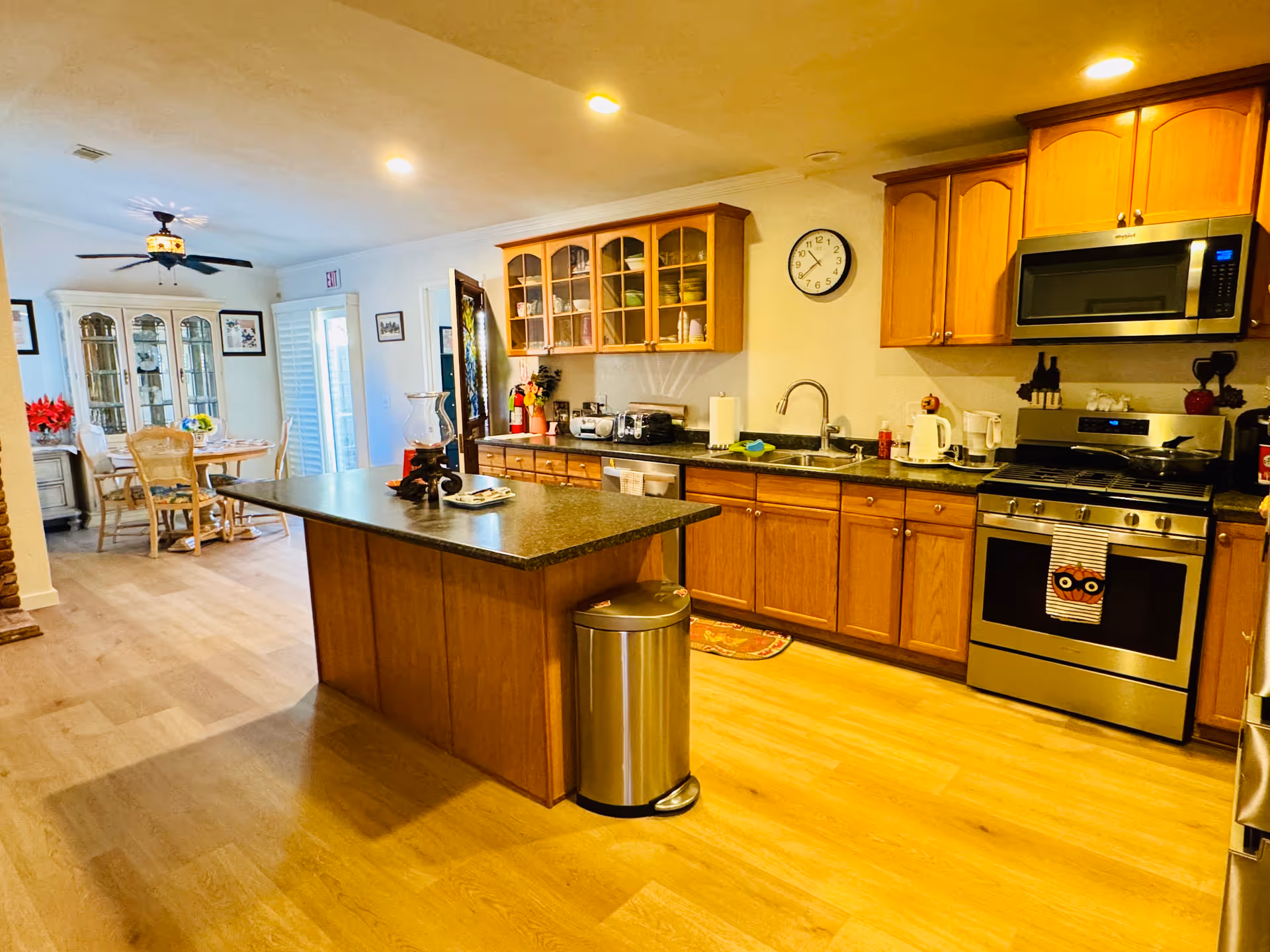 A bright kitchen with wooden cabinets, a central island with a dark countertop, stainless steel appliances including a stove and microwave, and a sink. In the background, there is a dining area with a table and chairs, a ceiling fan, and a glass-front cabinet.