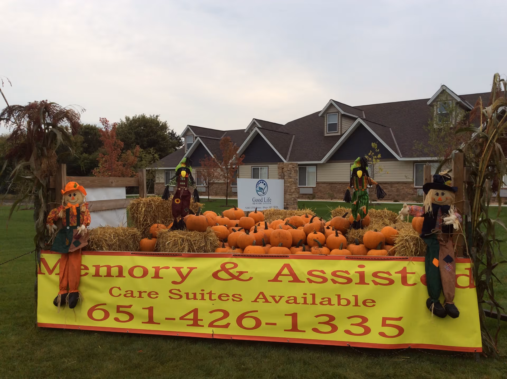 Pumpkin and hay bale display with scarecrows and a yellow banner advertising memory and assisted care in front of a Good Life Senior Living building.