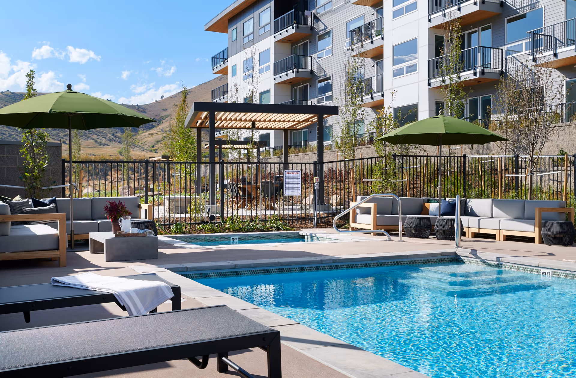 Outdoor pool area at a senior living facility with clear blue water, surrounded by lounge chairs with a towel, cushioned seating under green umbrellas, and a pergola with a table and chairs. The background shows a multi-story residential building and hills under a blue sky with some clouds.