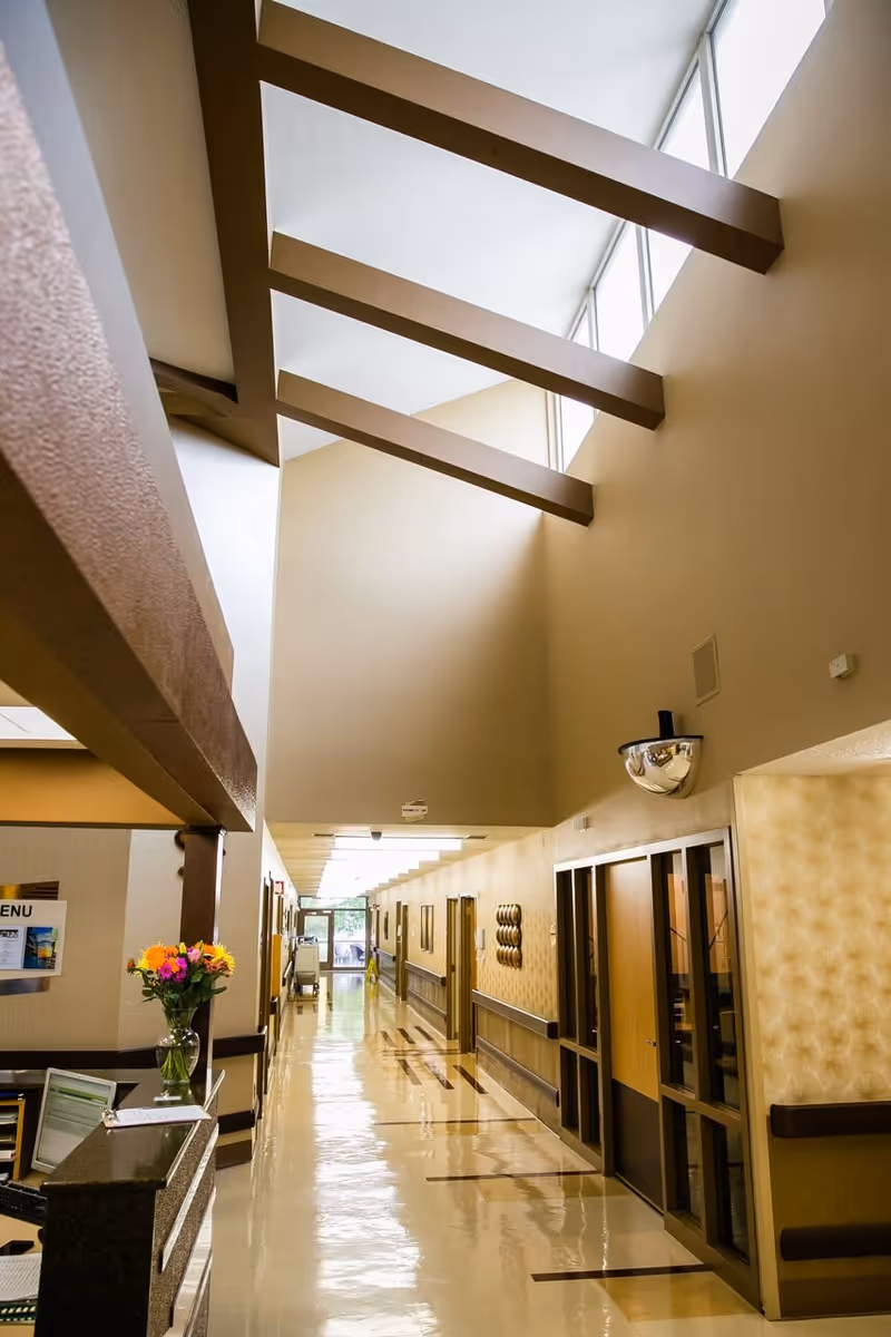 Well-lit interior hallway with a high skylit ceiling, reception desk with flowers and a polished floor leading to resident rooms.