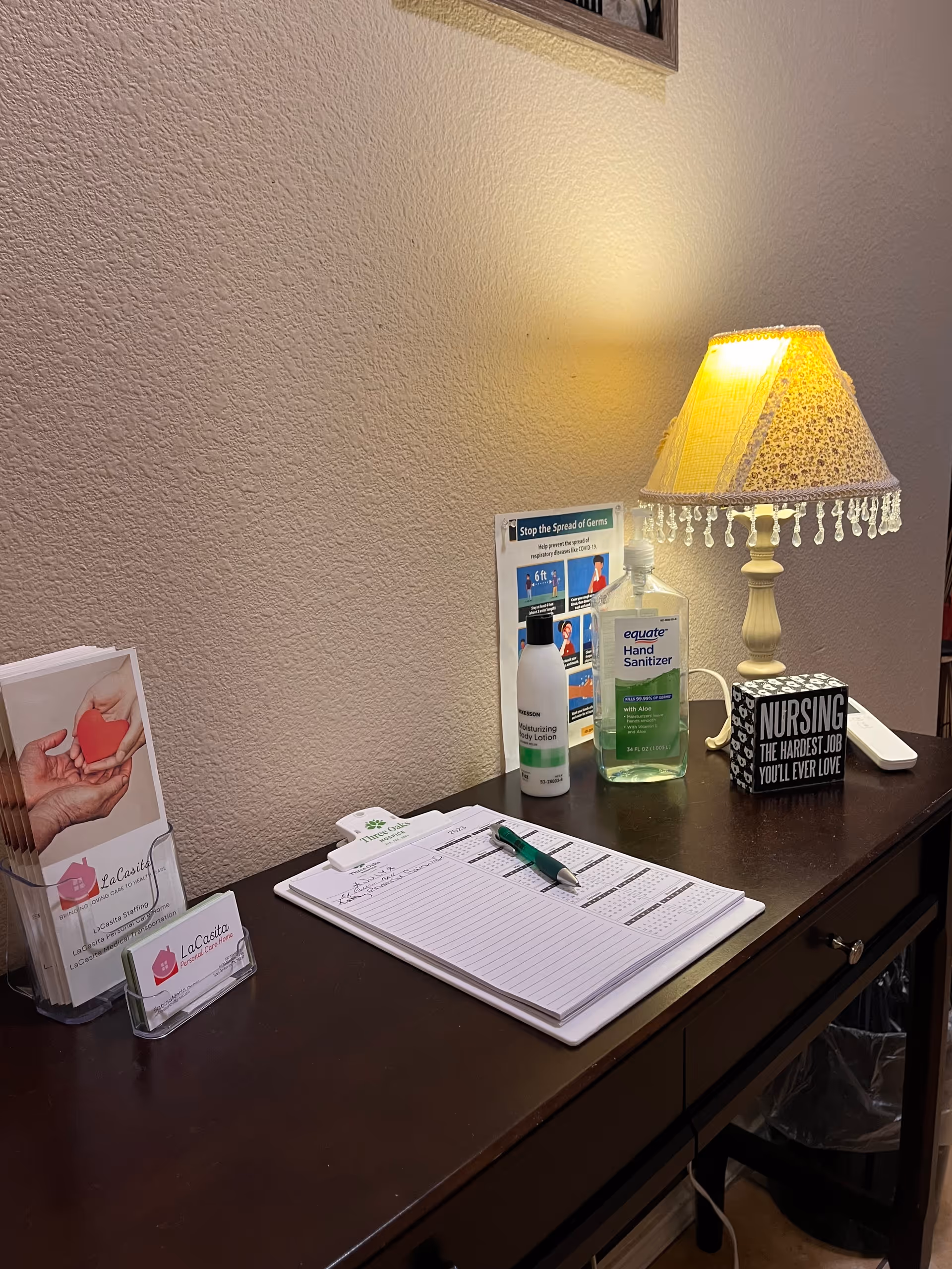 A dark wooden desk with a lamp, sanitizer, brochures, and forms in a care home office.