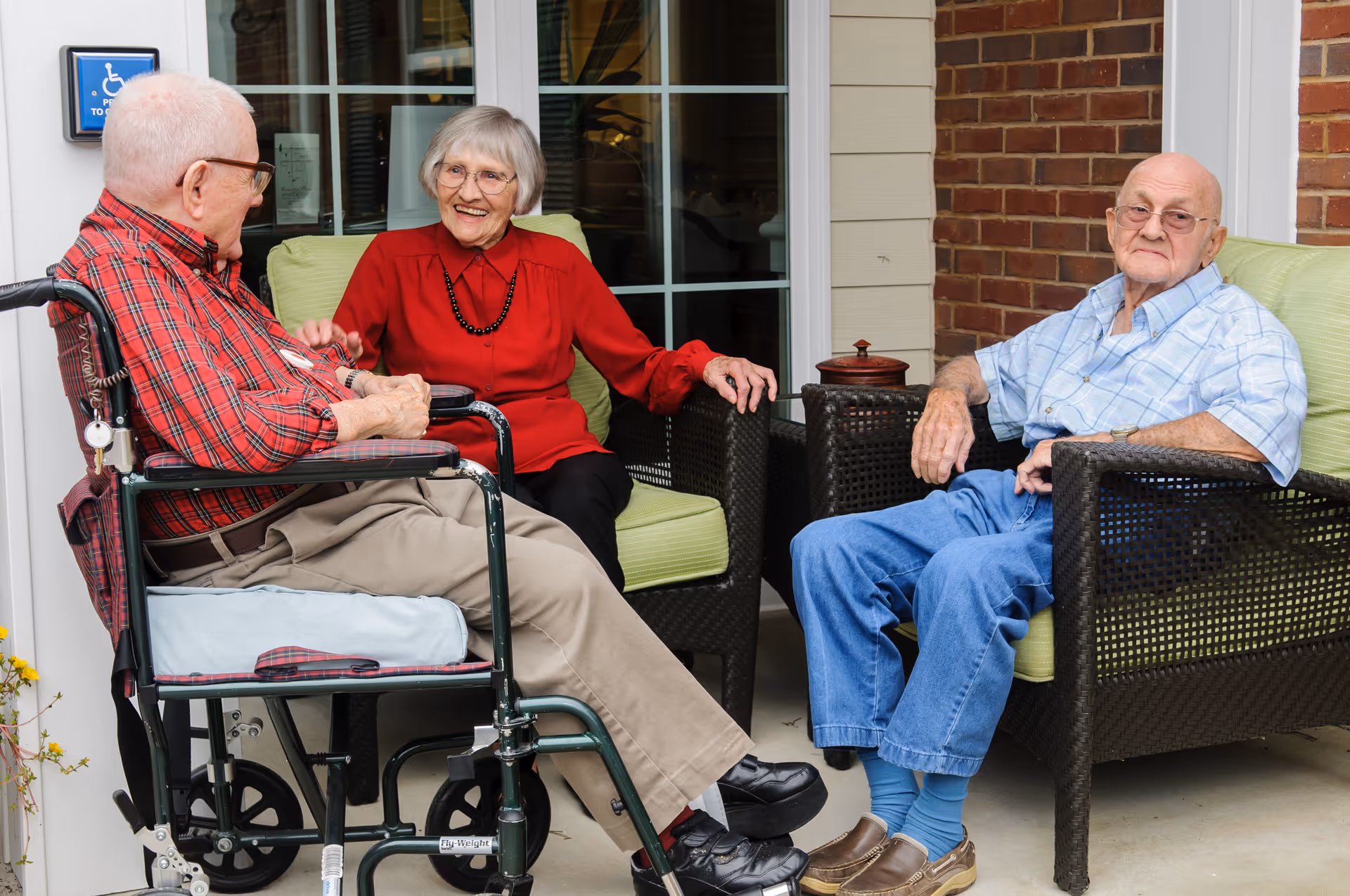 Three elderly individuals sitting and conversing on a patio outside a building. One man is in a wheelchair wearing a red plaid shirt and beige pants, a woman in a red blouse and black pants is seated on a green cushioned chair, and another man in a blue plaid shirt and blue pants is seated on a similar chair. The background shows a window and a brick wall.