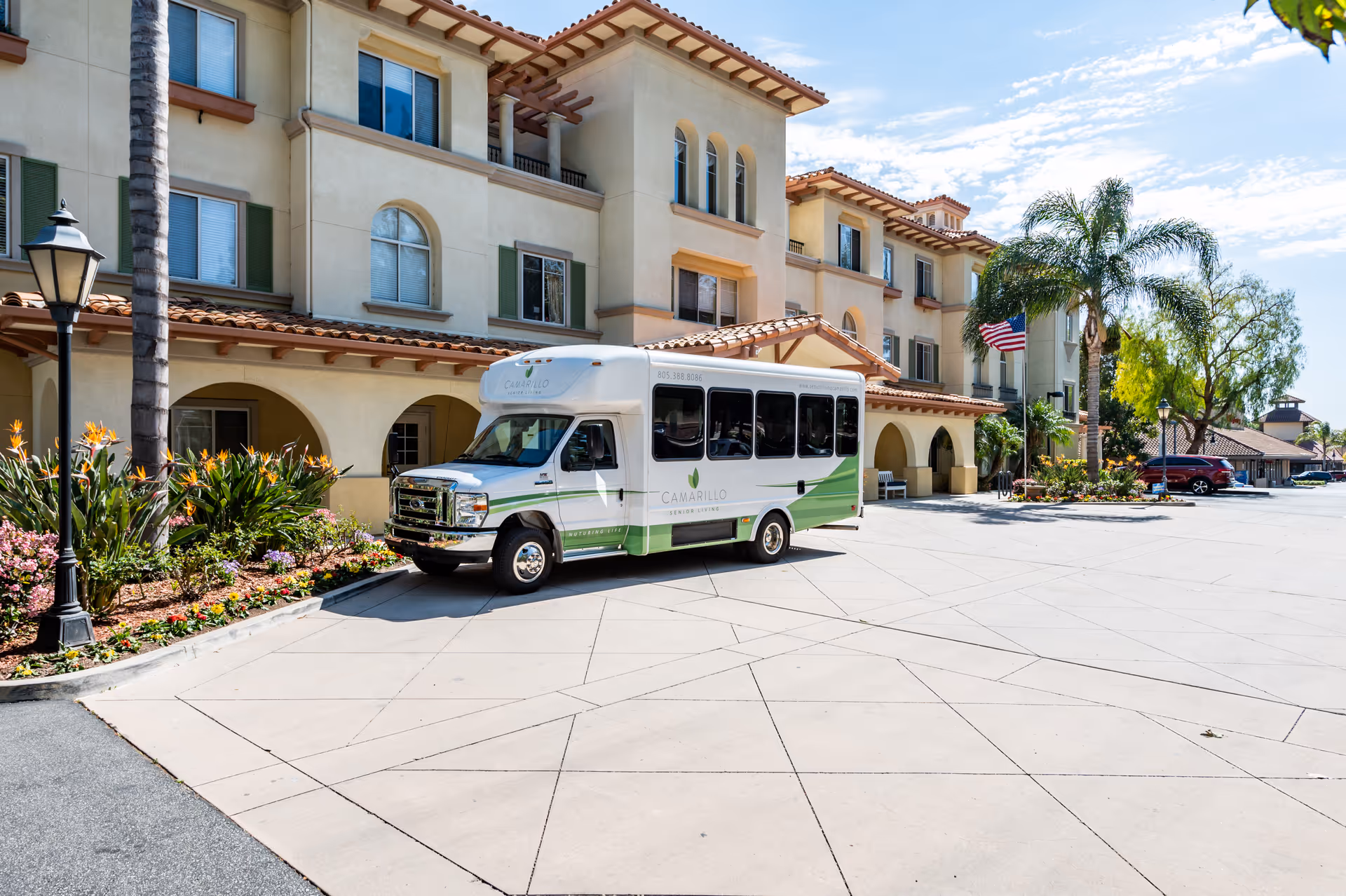 Exterior view of Camarillo Senior Living facility with a white and green shuttle bus parked in front. The building has a beige facade with arched windows and a tiled roof. There are palm trees, flowers, and an American flag near the entrance under a clear blue sky.