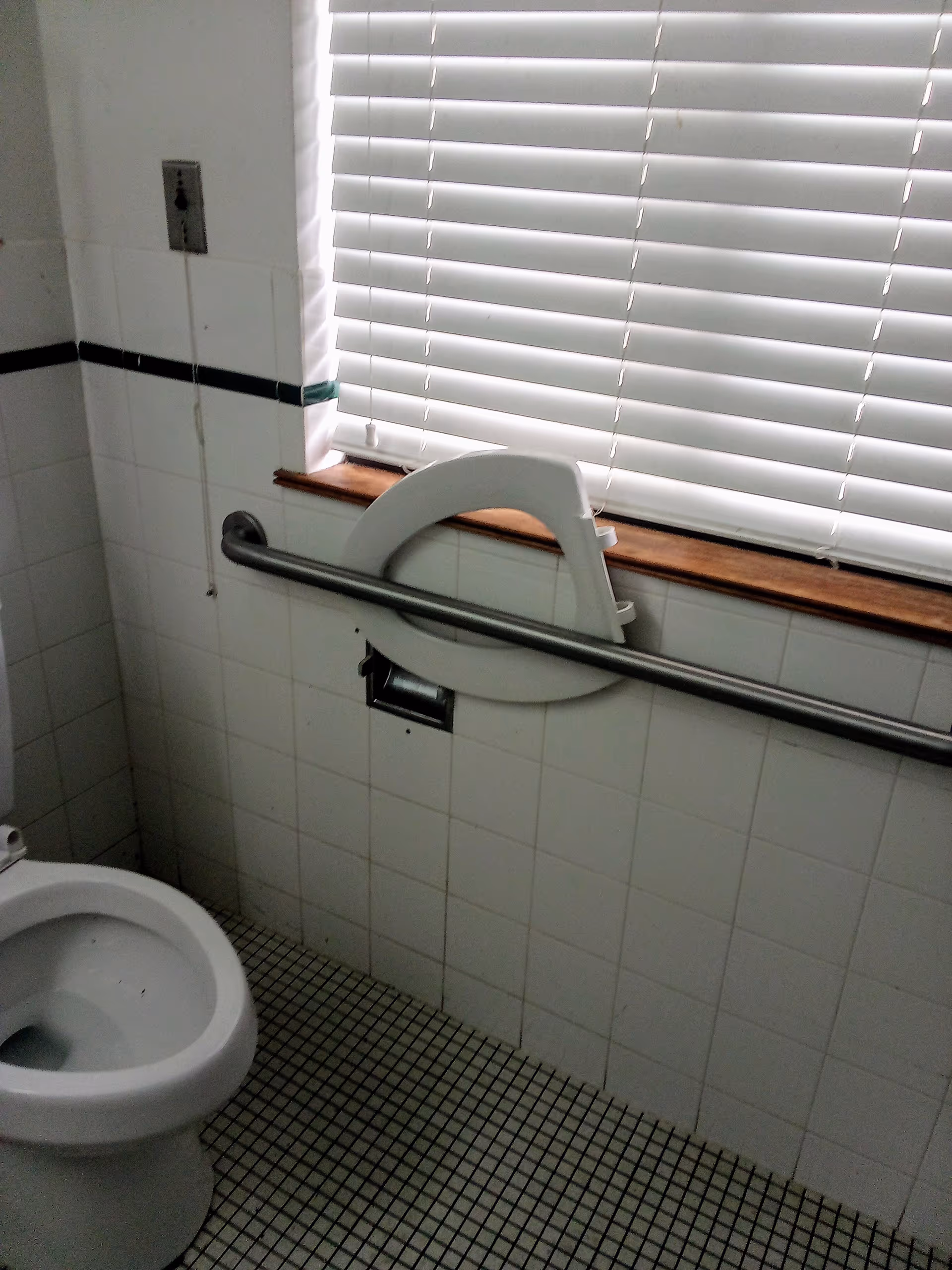 Accessible bathroom with tiled walls, toilet, grab bar, and blinds over a window.