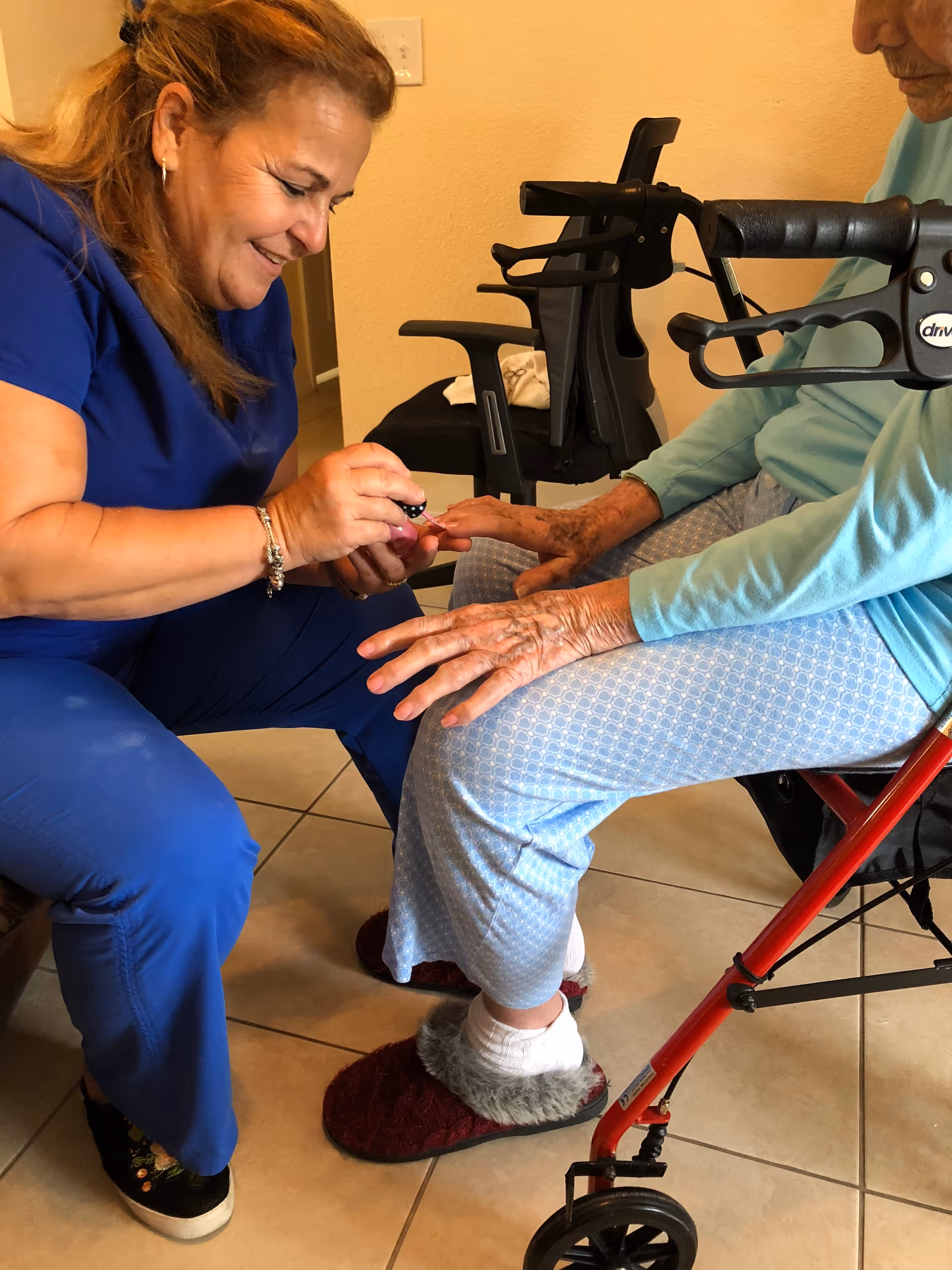 A caregiver in blue scrubs is painting the fingernails of an elderly person seated on a red walker. The elderly person is wearing light blue patterned pants, a light blue long-sleeve shirt, white socks, and red slippers. The scene takes place indoors on a tiled floor.