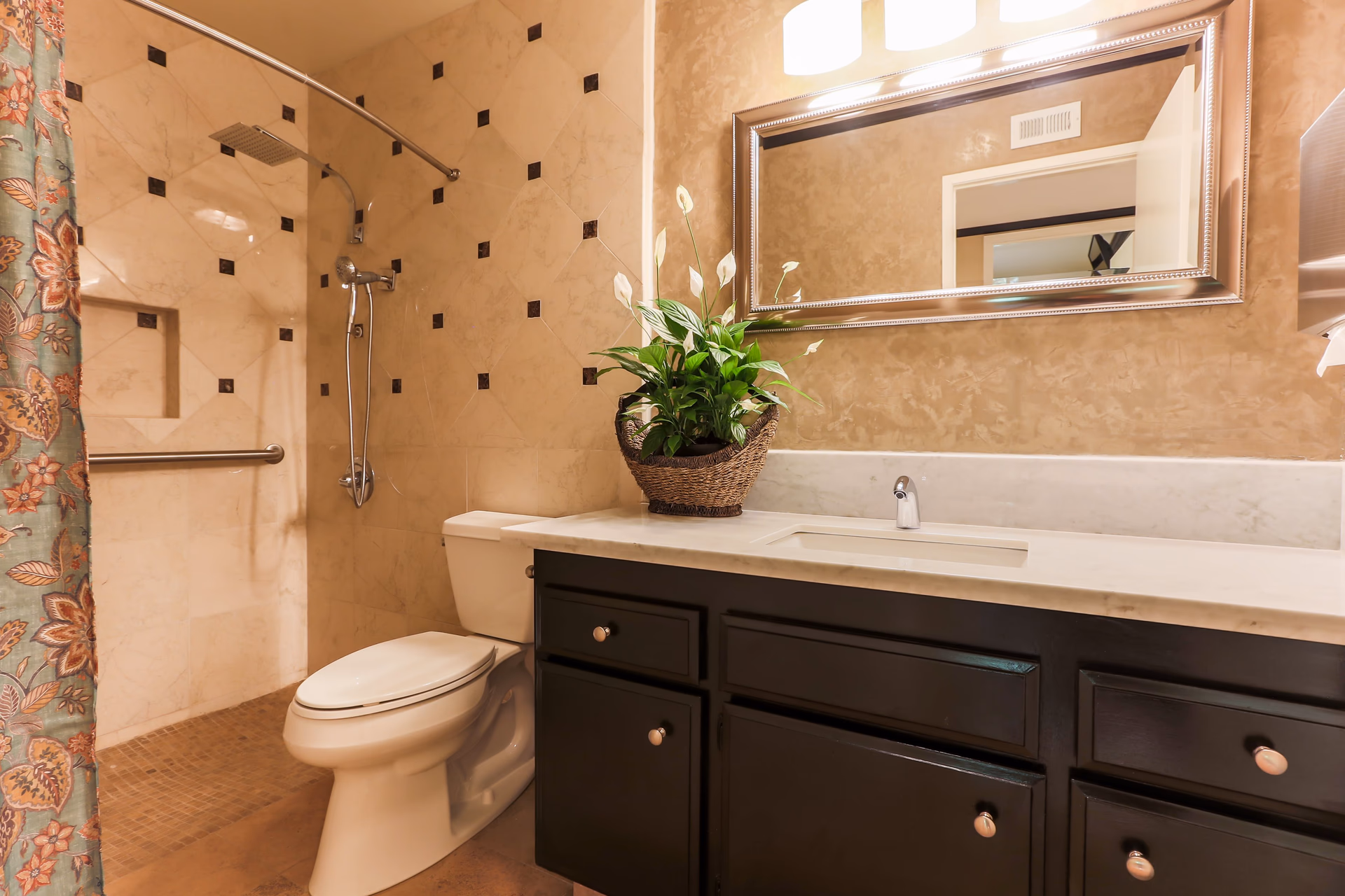 A bathroom with beige tiled walls featuring small dark square accents, a white toilet, a shower area with a curved shower rod and floral shower curtain, a dark wood vanity with a white countertop and an integrated sink, a large rectangular mirror with a silver frame above the vanity, and a potted green plant in a woven basket on the countertop.