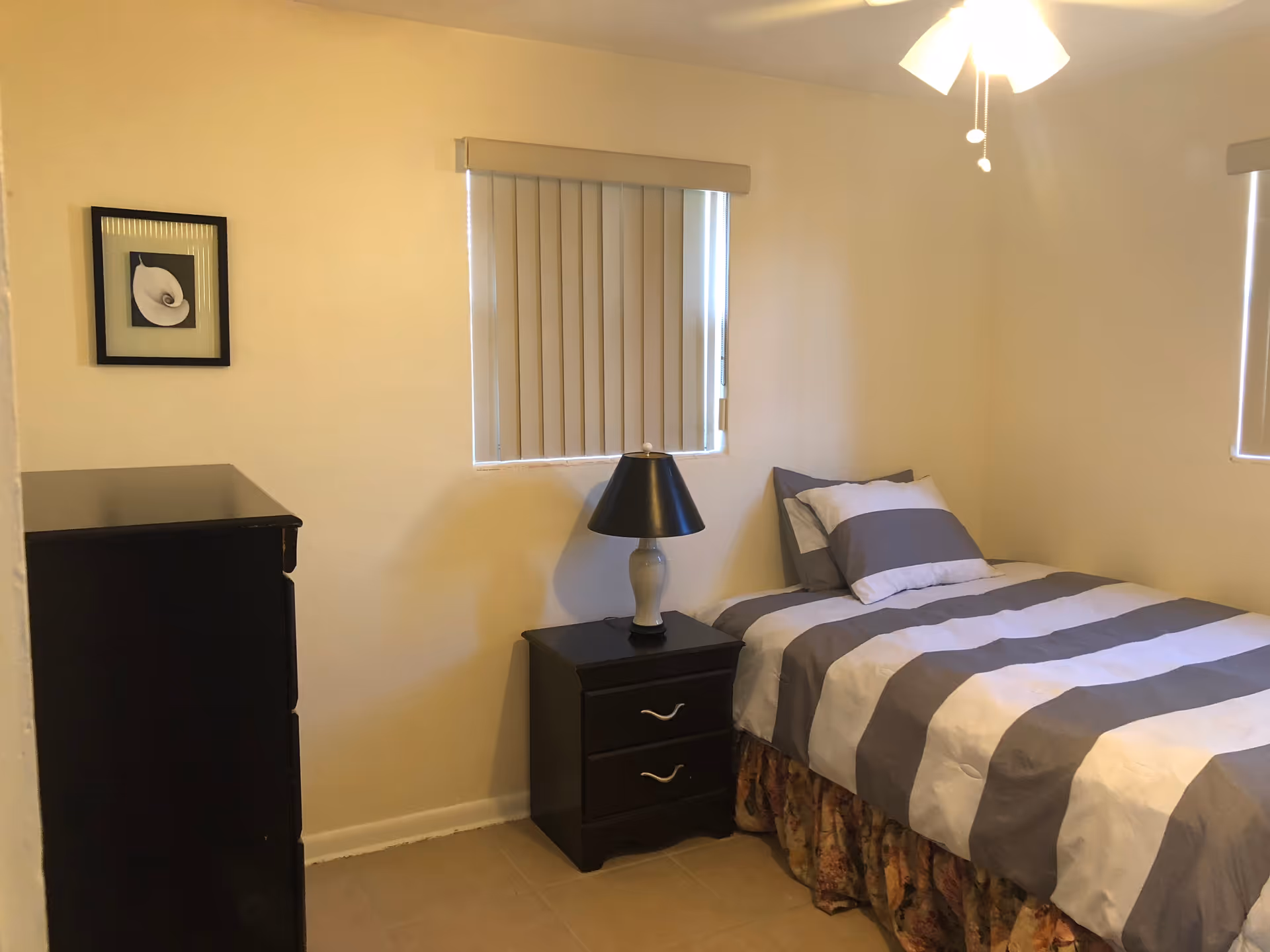 A small bedroom in a facility with a striped comforter, nightstand, lamp, dresser, and blinds.