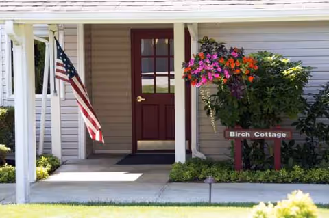 Front entrance of Birch Cottage at a senior living facility, with flag and hanging flowers.