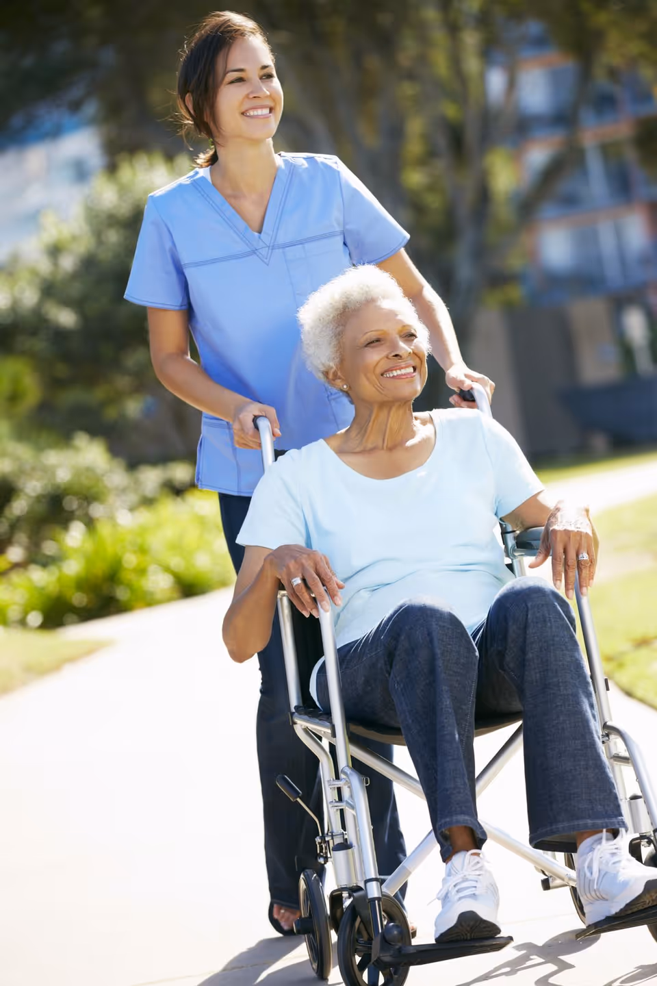 Caregiver in blue scrubs pushes elderly woman in a wheelchair along a sunny outdoor path.