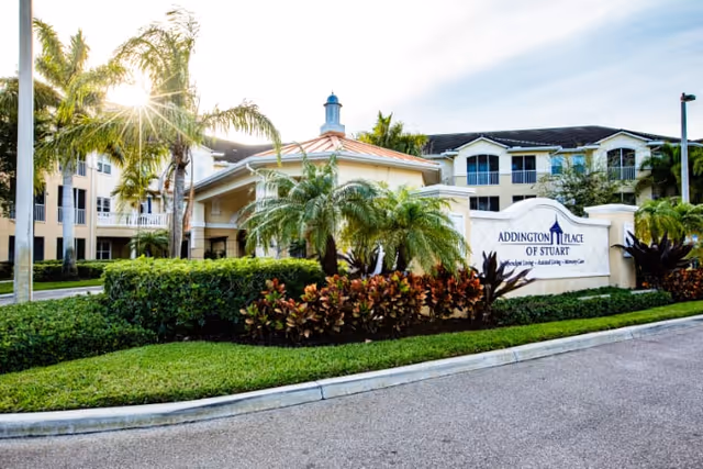 Exterior view of Addington Place of Stuart senior living facility with landscaped greenery, palm trees, and a sign displaying the facility name. The building is multi-story with balconies and a light-colored facade under a sunny sky.