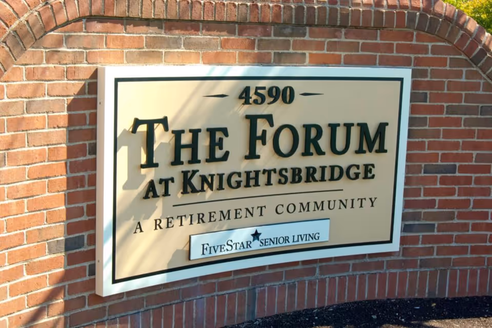 A large beige sign mounted on a red brick wall with the text '4590 The Forum at Knightsbridge A Retirement Community Five Star Senior Living'.