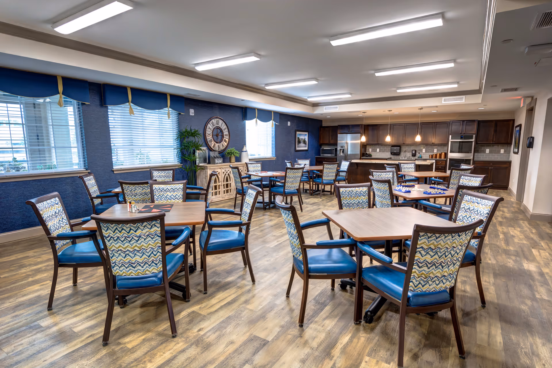A spacious senior living dining area with multiple wooden tables and chairs featuring blue patterned upholstery. The room has large windows with blue valances, a wooden floor, and a kitchen area with dark wood cabinets and stainless steel appliances in the background. The walls are painted blue and decorated with a large clock and plants.