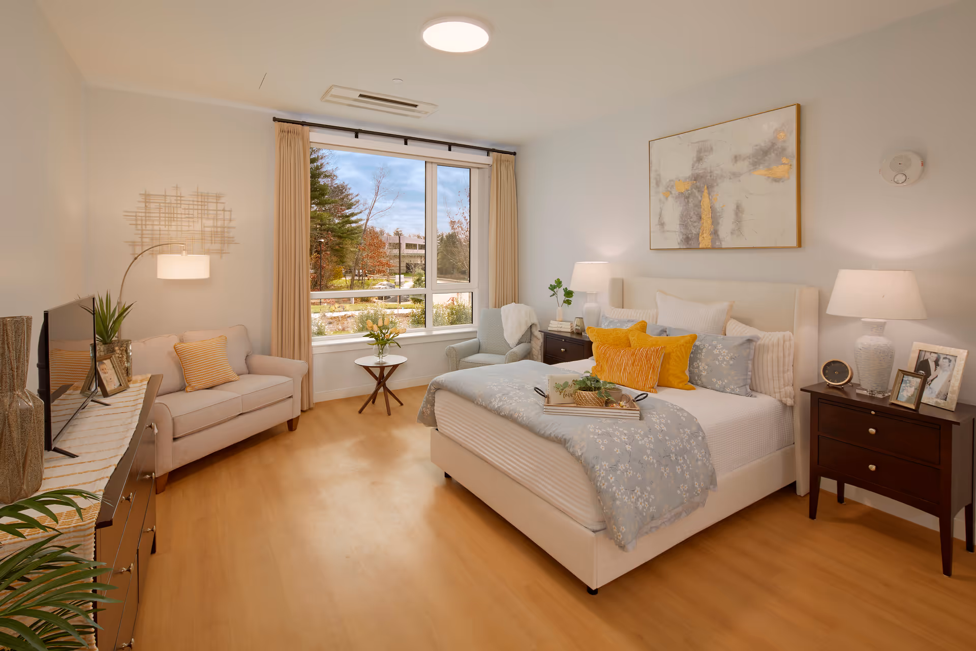 Bright bedroom in a senior living facility with a bed, seating area, and large window.