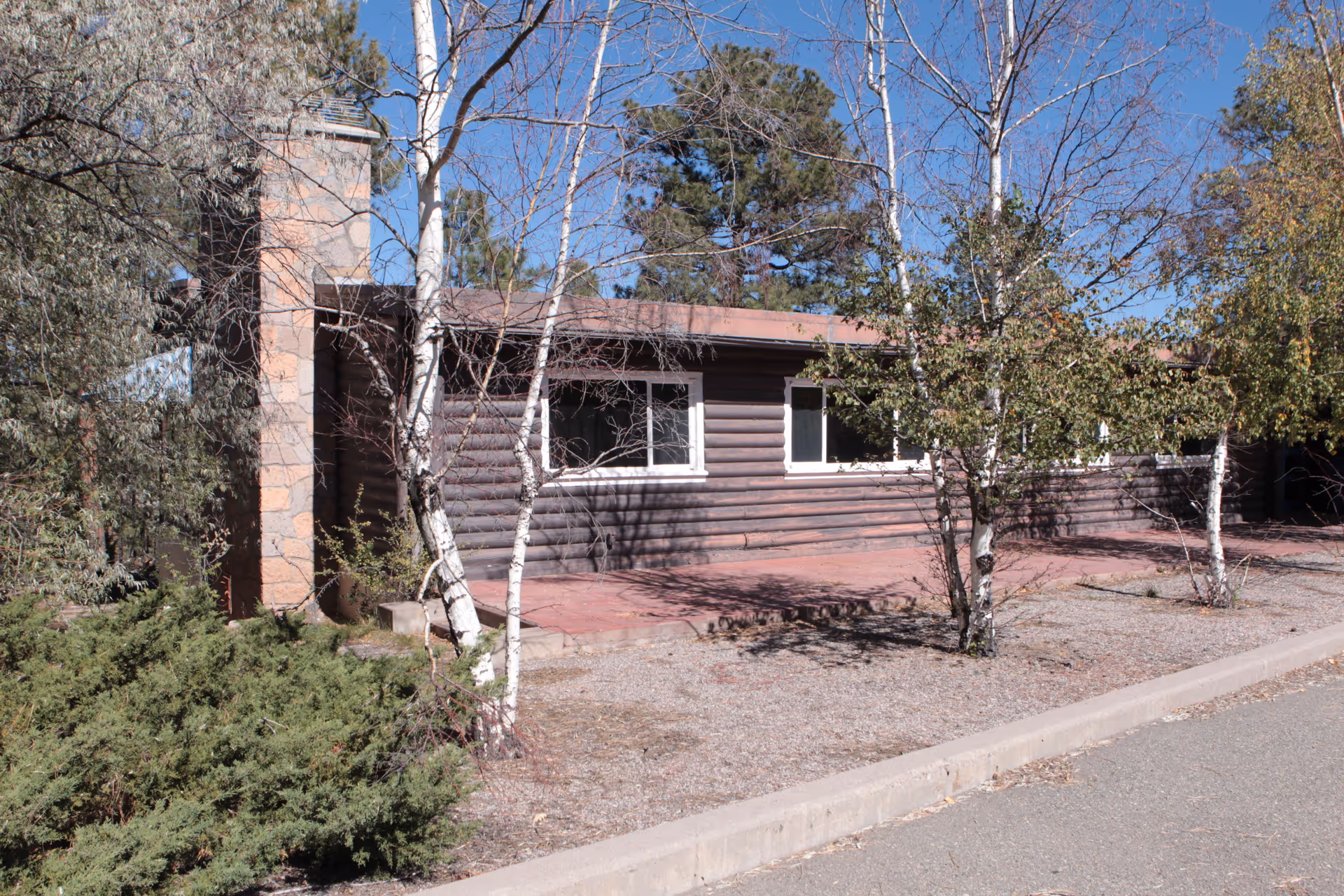 A single-story brown wooden building exterior with trees and a sidewalk.
