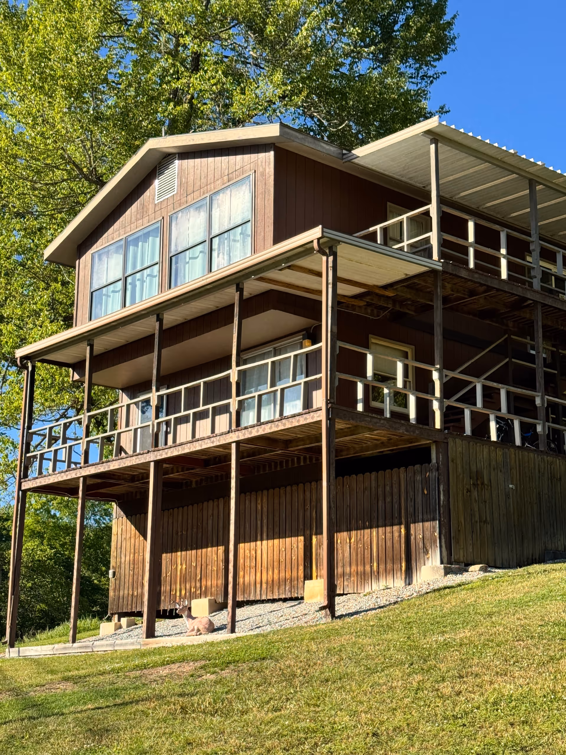 Two-story wooden house on stilts with wraparound balcony beside green trees.