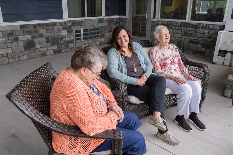 Three elderly women sit on wicker chairs on a BeeHive Homes patio.
